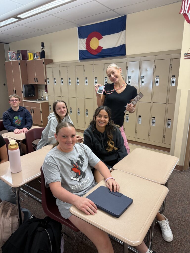 Students sit at desks in a classroom with Colorado's flag in the background. One student holds a badge and pamphlet.