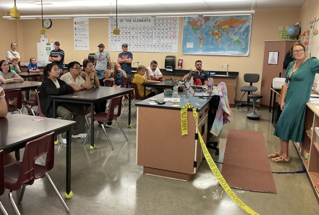 A diverse group of people attentively listens in a classroom with science materials and a world map on the wall.
