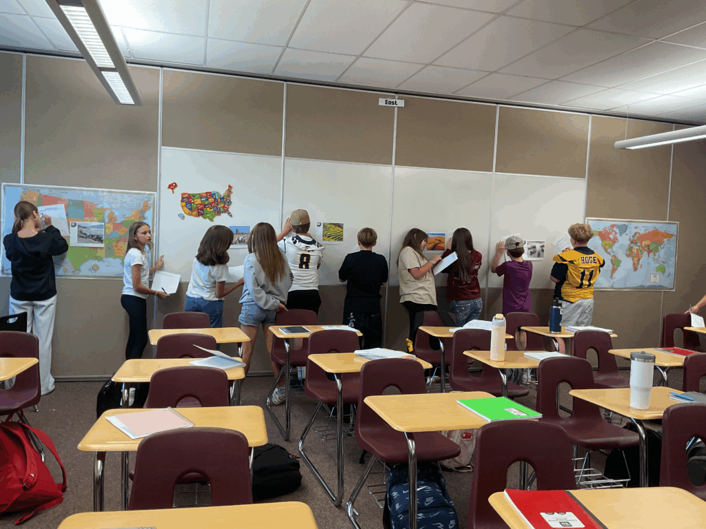 Students in a classroom are engaged in an activity, reviewing maps and images displayed on the walls.