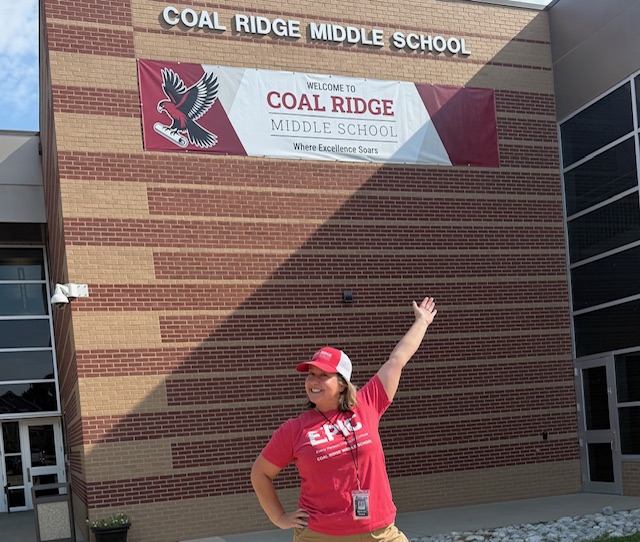 A smiling woman in a red shirt stands outside Coal Ridge Middle School, pointing at a welcome banner.