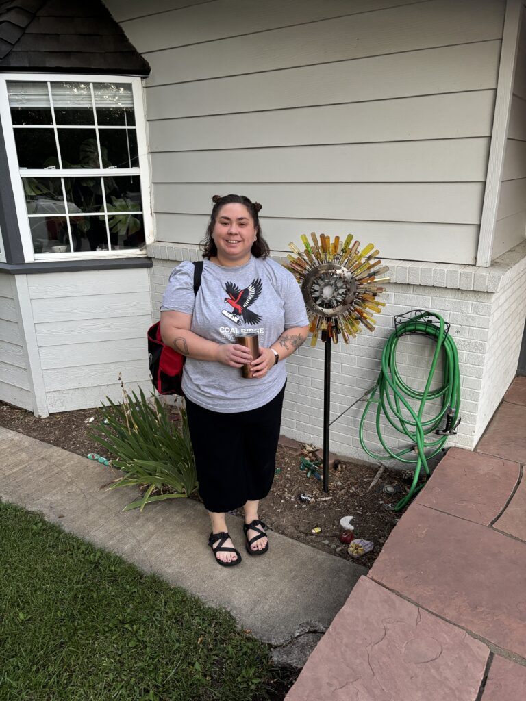 A woman stands outside a house holding a drink, next to a colorful garden decoration and green garden hose.