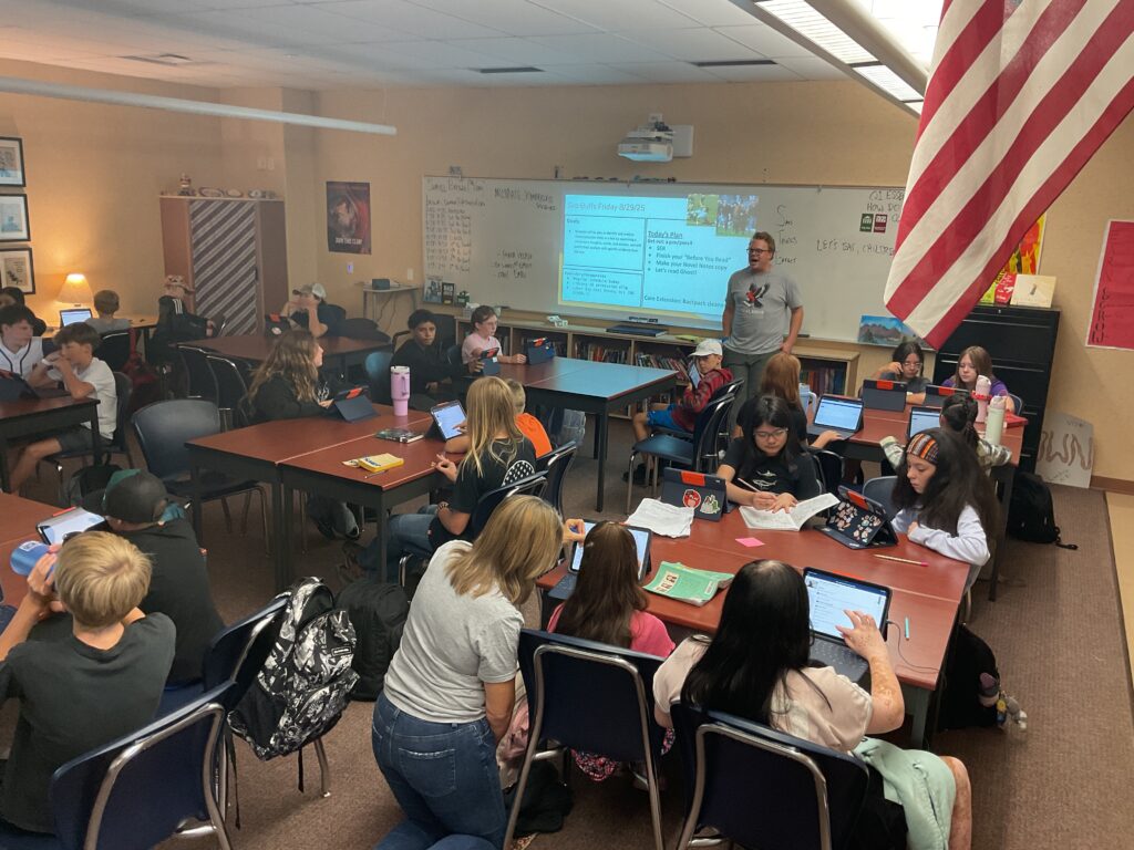 A classroom filled with students using tablets, with a teacher presenting information on a screen. An American flag is visible.