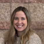 Smiling woman with long hair wearing a beige sweater, posed against a textured brown wall.