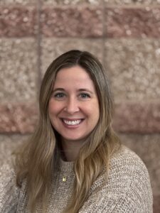 Smiling woman with long hair wearing a beige sweater, posed against a textured brown wall.
