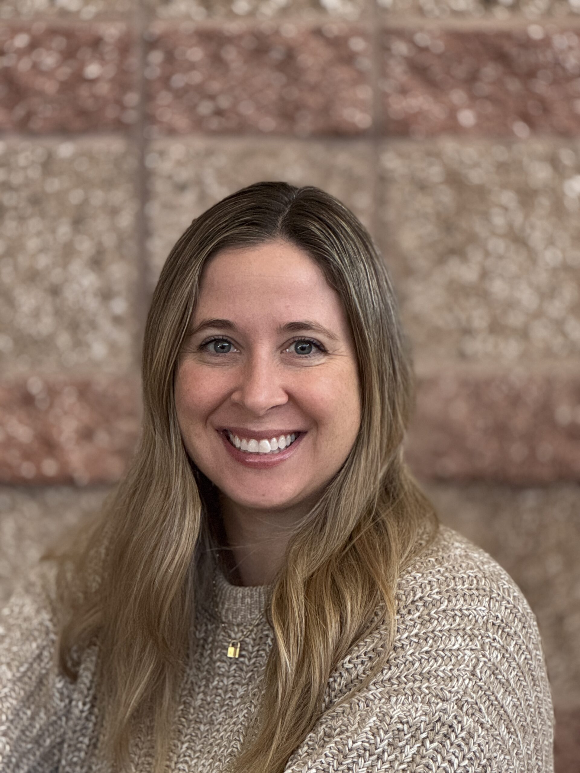Smiling woman with long hair wearing a beige sweater, posed against a textured brown wall.