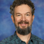 Smiling man with curly hair and a beard, wearing a patterned shirt, against a blue background.