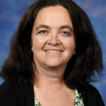 Smiling woman with dark, wavy hair, wearing a floral blouse and black cardigan, set against a blue background.