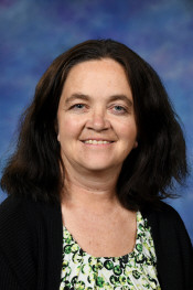Smiling woman with dark, wavy hair, wearing a floral blouse and black cardigan, set against a blue background.