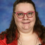 A smiling woman with long brown hair, wearing pink glasses and a red jacket over a polka dot dress, against a blue background.