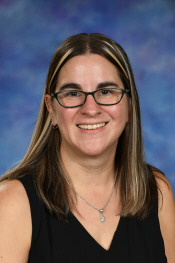 A smiling woman with long brown hair and glasses, wearing a black top, against a blue background.