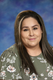 Smiling woman with long dark hair, wearing a floral green blouse, against a blue background.