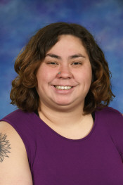 A smiling person with curly brown hair, wearing a purple top, against a blue background.