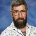 A man with a beard and glasses, wearing a patterned shirt, poses against a blue background.