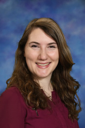 A young woman with long, wavy brown hair smiles warmly against a blue and purple background. She wears a burgundy shirt.