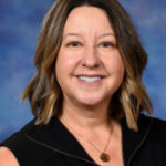 A smiling woman with shoulder-length brown hair, wearing a black vest and a necklace, against a blue background.