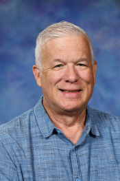 A smiling older man with short gray hair, wearing a blue button-up shirt, against a blue background.