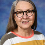 A smiling woman with long gray hair and glasses wears a striped shirt, posing against a blue background.