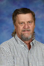 A man with a beard and mullet hairstyle, wearing a checkered shirt, smiles against a blue background.