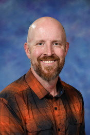 Smiling man with a bald head and a beard, wearing an orange and black checkered shirt, against a blue background.