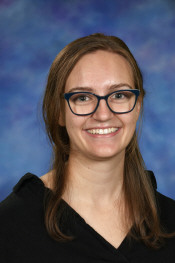 A smiling woman with glasses and brown hair, wearing a black top, against a blue and purple background.
