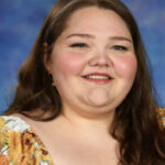 A smiling young woman with long brown hair, wearing a floral blouse, poses against a blue background.