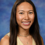 Una joven de pelo largo y oscuro sonríe con calidez, vestida con un top amarillo claro y un collar de cuentas, sobre un fondo azul.