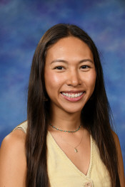 A young woman with long dark hair smiles warmly, wearing a light yellow top and a beaded necklace, against a blue background.