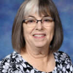 A smiling woman with shoulder-length hair and glasses, wearing a black and white floral top, against a blue background.