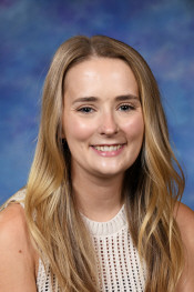A smiling young woman with long, wavy hair, wearing a sleeveless top, against a blue and purple background.