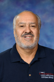 A smiling man with a beard, wearing a dark polo shirt with a logo, set against a blue background.