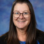 Smiling woman with long brown hair and glasses, wearing a blue shirt against a blurred blue background.