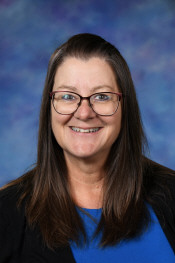 Smiling woman with long brown hair and glasses, wearing a blue shirt against a blurred blue background.