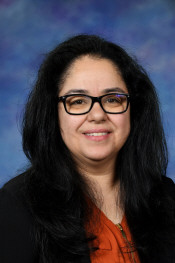 A woman with long black hair and glasses smiles against a blue and purple background. She wears an orange top.