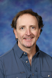 Smiling man with curly hair wearing a blue shirt, set against a soft blue background.