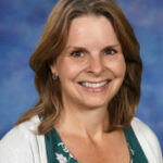 Smiling woman with shoulder-length brown hair, wearing a green floral top and white cardigan, against a blue background.