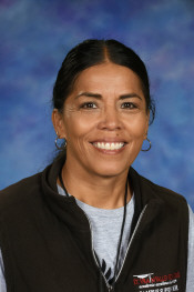 A smiling woman with long dark hair, wearing a brown vest over a gray shirt, set against a blue background.
