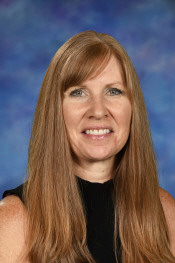 Portrait of a woman with long brown hair, smiling, against a blue and purple background. She wears a black top.