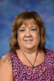 A woman with brown, wavy hair smiles at the camera, wearing a pink patterned top and a necklace. Background is blue.
