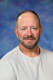 A middle-aged man with a beard smiles at the camera, wearing a light shirt against a blue background.