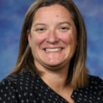 A smiling woman with shoulder-length brown hair, wearing a black blouse, against a blue background.