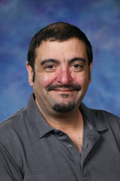 A man with dark hair and a mustache smiles, wearing a gray shirt against a blue background.