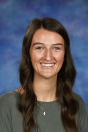 A smiling young woman with long brown hair, wearing a green top, against a blue and purple background.