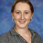 Smiling young woman with long hair, wearing a checkered shirt and hoop earrings, against a blue background.