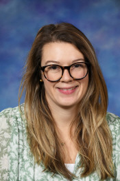 A smiling woman with long, wavy hair, wearing glasses and a floral blouse, poses against a blue background.