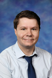 A smiling man in a light blue shirt and dark tie poses against a blue gradient background.