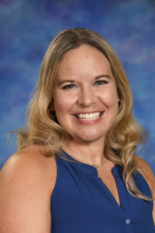 A smiling woman with long, wavy blonde hair, wearing a blue shirt, against a colorful backdrop.