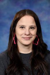 A woman with long dark hair and pink flower-shaped earrings smiles against a blue background.