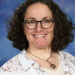 A smiling woman with curly hair and glasses, wearing a patterned shirt, poses against a blue background.