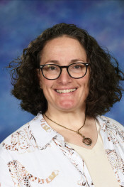 A smiling woman with curly hair and glasses, wearing a patterned shirt, poses against a blue background.