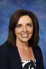 Smiling woman with shoulder-length brown hair, wearing a dark cardigan over a patterned top, against a blue background.